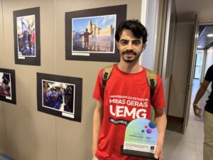 Lucas segura o troféu e está posicionado perto da fotografia que fez, colada em uma parede do CRP-MG.